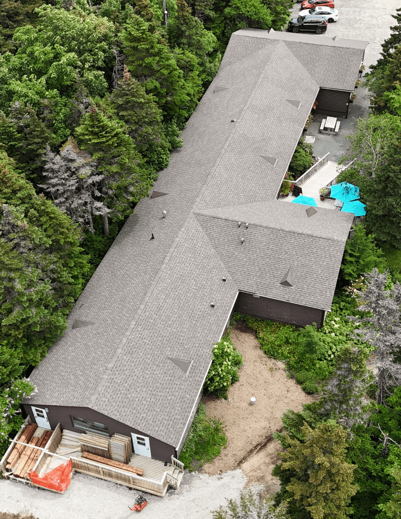 Aerial view of a T-shaped building with a shingled roof surrounded by trees, featuring a pool area