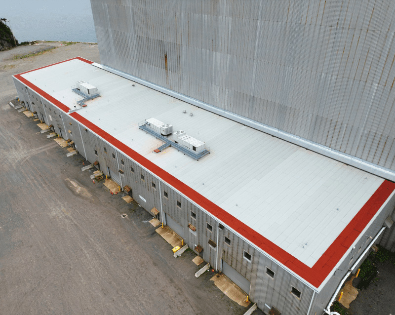 Aerial view of industrial buildings with red trim near a forested hillside and a water body
