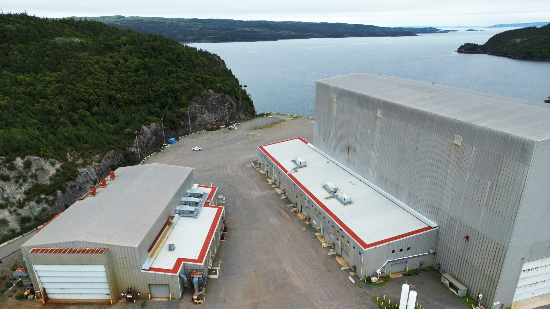 Aerial view of industrial buildings with red trim near a forested hillside and a water body